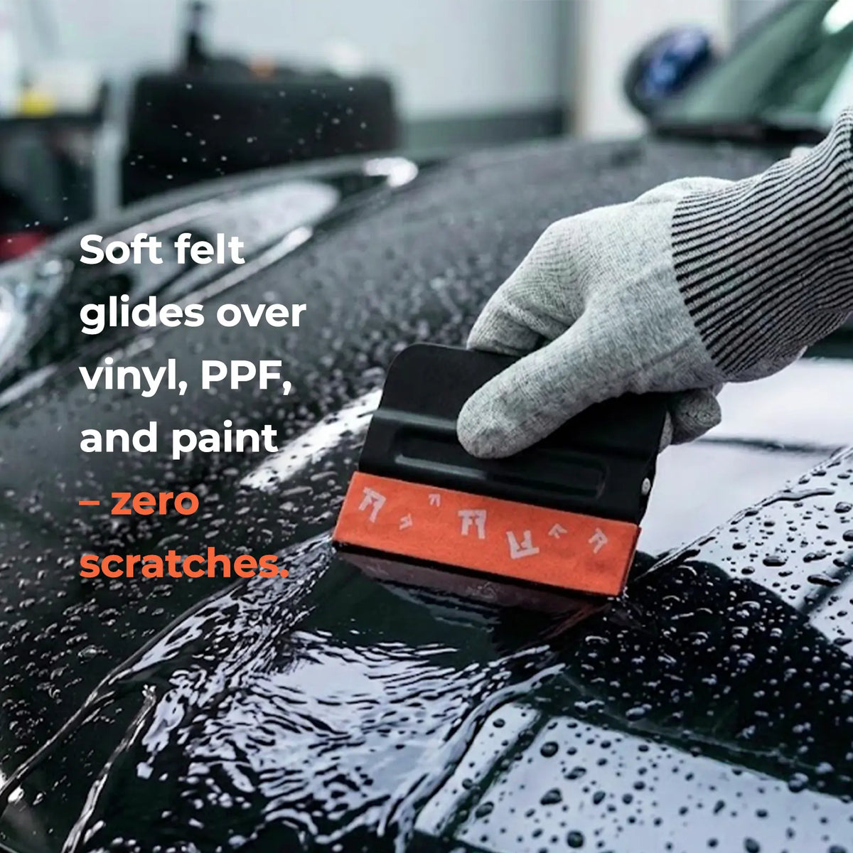 A gloved hand uses a black and orange felt car scratch remover on a wet black car surface with water droplets. White and orange text on the left reads, 'Soft felt glides over vinyl, PPF, and paint - zero scratches.