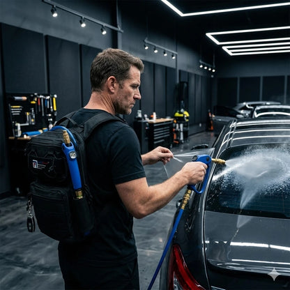 A man in a black t-shirt and black backpack sprays a dark gray car with a hose in a modern garage setting.