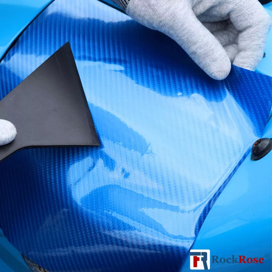 Person applying blue car wrap to a vehicle with a black tool, wearing gray gloves. on blue carbon fiber