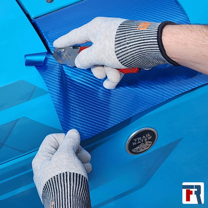 Person cutting blue carbon vinyl wrap with a tool on a blue surface, with a brand logo in the corner.