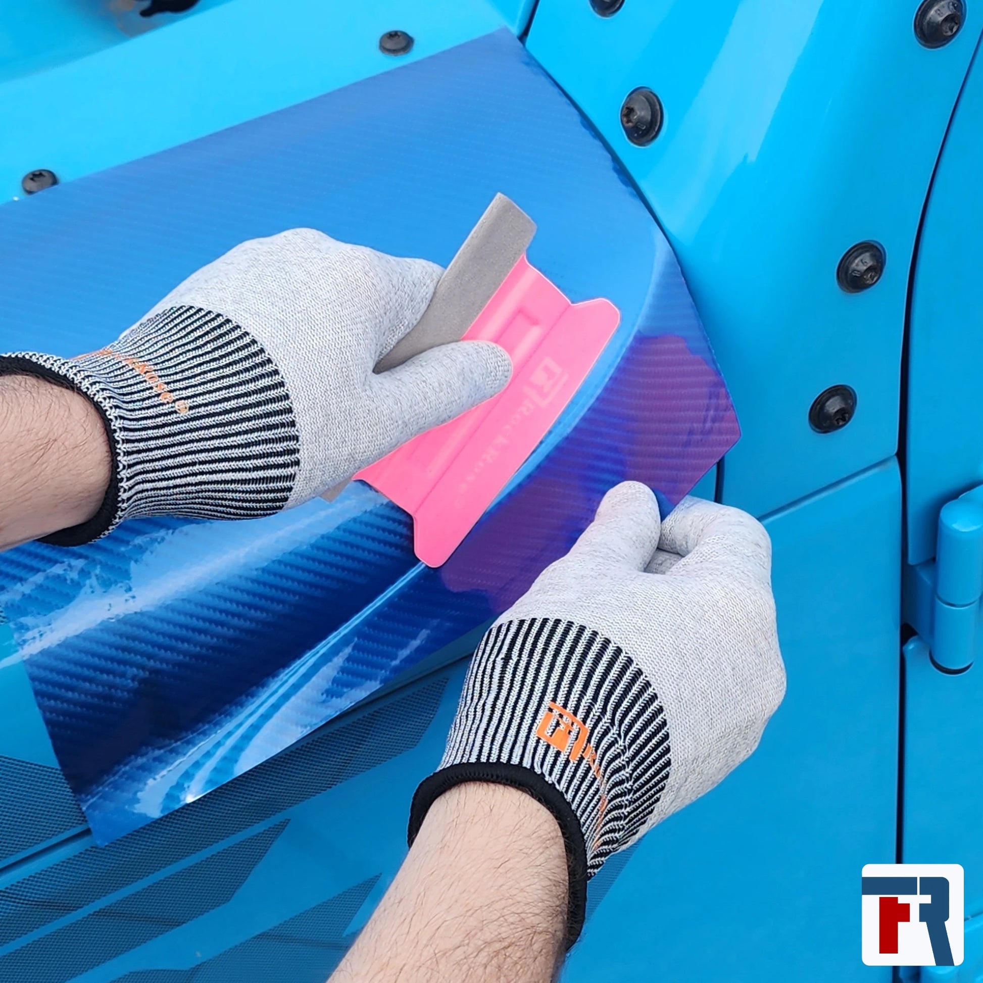 Person applying blue carbon vinyl wrap to a blue vehicle panel with a pink squeegee 
