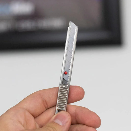 A hand holding a silver utility knife with a white blade and a small red indicator dot, shown against a blurred indoor background.