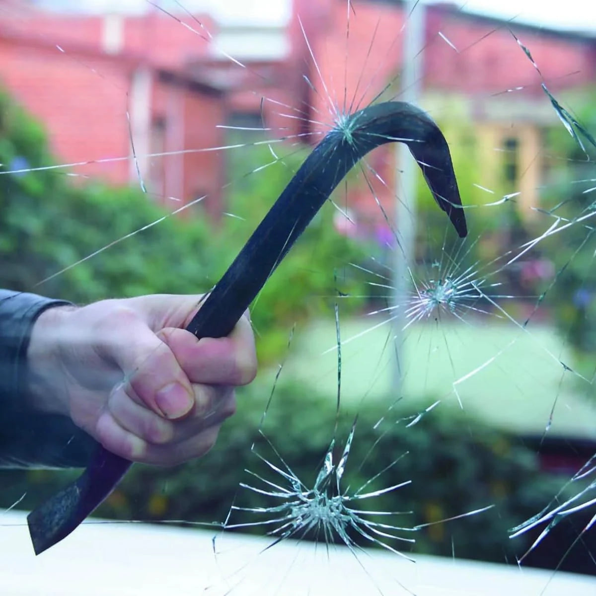 A hand holding a black crowbar strikes and shatters a glass pane with multiple cracks radiating from the impact point, with a blurred outdoor background of greenery and a red brick building.