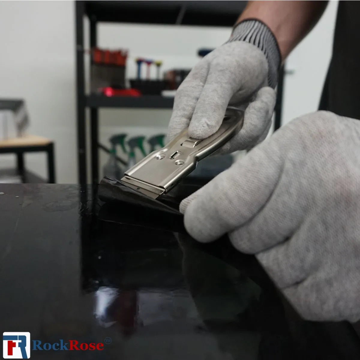 Person wearing gloves using a Steel Razor Scraper on a black surface with 'RockRose' branding.